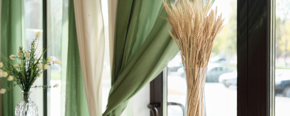 partial view of stylish interior of cafe or terrace with natural decor. ear of wheat in a glass vase on light wooden table, green curtains and flowers. window view to a street